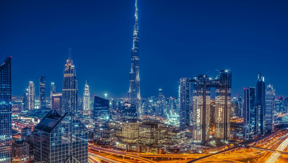 Night view of Dubai city skyline with Burj Khalifa, illuminated skyscrapers, and busy highways with flowing traffic lights.