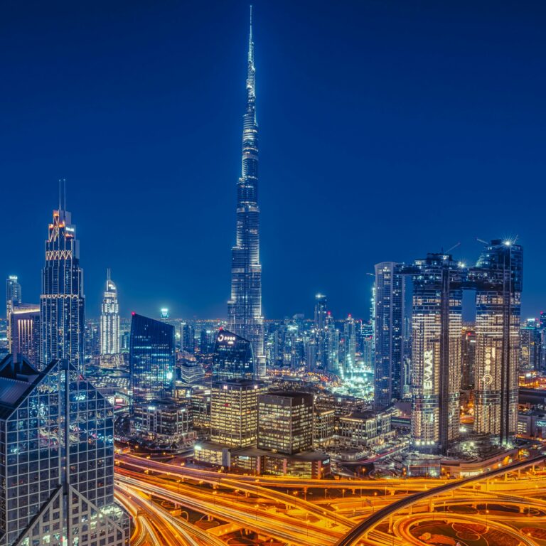 Night view of Dubai city skyline with Burj Khalifa, illuminated skyscrapers, and busy highways with flowing traffic lights.