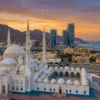 Aerial view of the Sheikh Zayed Grand Mosque during a Fujairah City Tour, with white domes and minarets framed by mountains, modern buildings, and the coastline at sunset.