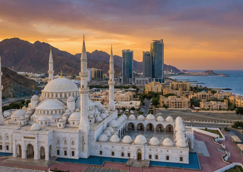Aerial view of the Sheikh Zayed Grand Mosque during a Fujairah City Tour, with white domes and minarets framed by mountains, modern buildings, and the coastline at sunset.