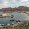 A scenic view resembling a Khorfakkan Tour, showcasing a grand mountain-side amphitheatre, sandy beach with visitors, turquoise coastline, and traditional boats along the shore against rugged rocky mountains.