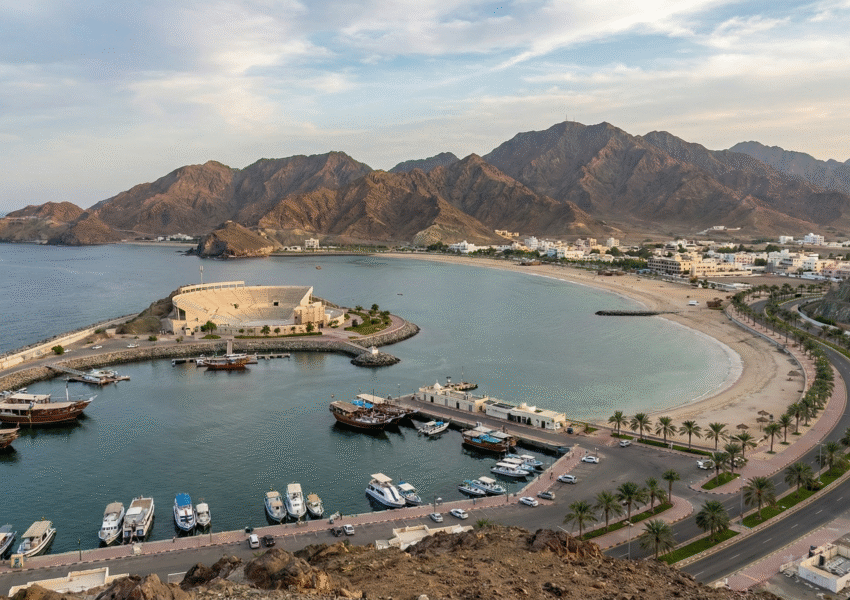 A scenic view resembling a Khorfakkan Tour, showcasing a grand mountain-side amphitheatre, sandy beach with visitors, turquoise coastline, and traditional boats along the shore against rugged rocky mountains.