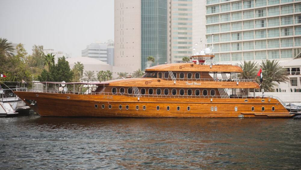 Luxury wooden dhow cruise Deira Creek sailing on Dubai Creek with modern city buildings in the background.