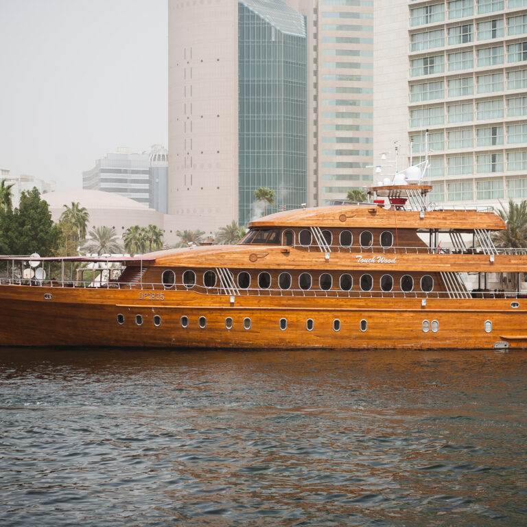 Luxury wooden dhow cruise Deira Creek sailing on Dubai Creek with modern city buildings in the background.
