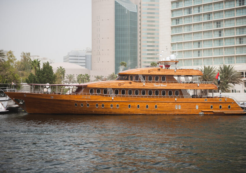 Luxury wooden dhow cruise Deira Creek sailing on Dubai Creek with modern city buildings in the background.