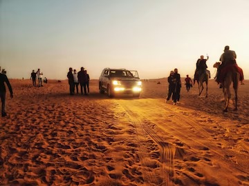 Dubai desert activities at sunset with a 4x4 vehicle driving through golden sand dunes, camel riders and visitors enjoying an evening desert safari experience.