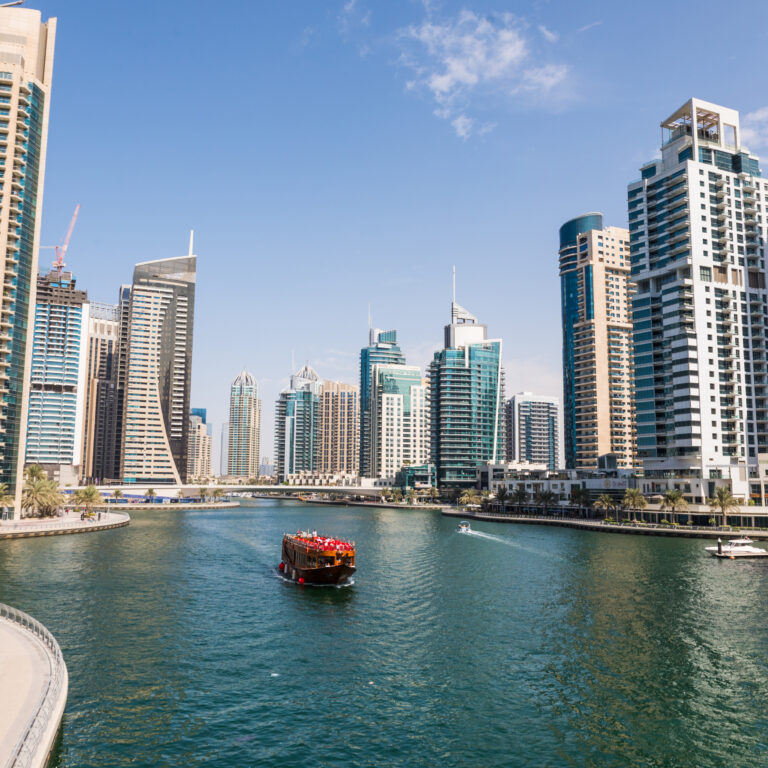 Traditional dhow cruise Dubai sailing through Dubai Marina canal with modern skyscrapers, waterfront promenade, and clear blue sky in the background.