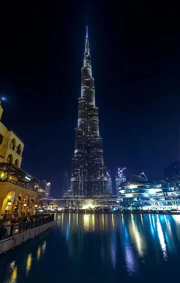 Dubai Night City Tour showcasing the illuminated Burj Khalifa reflecting on the waterfront, with modern buildings and vibrant city lights at night.
