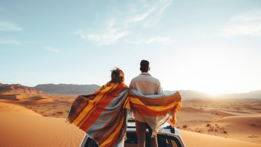 Couple standing on a 4x4 vehicle wrapped in blankets, overlooking golden sand dunes at sunset during a private desert safari.